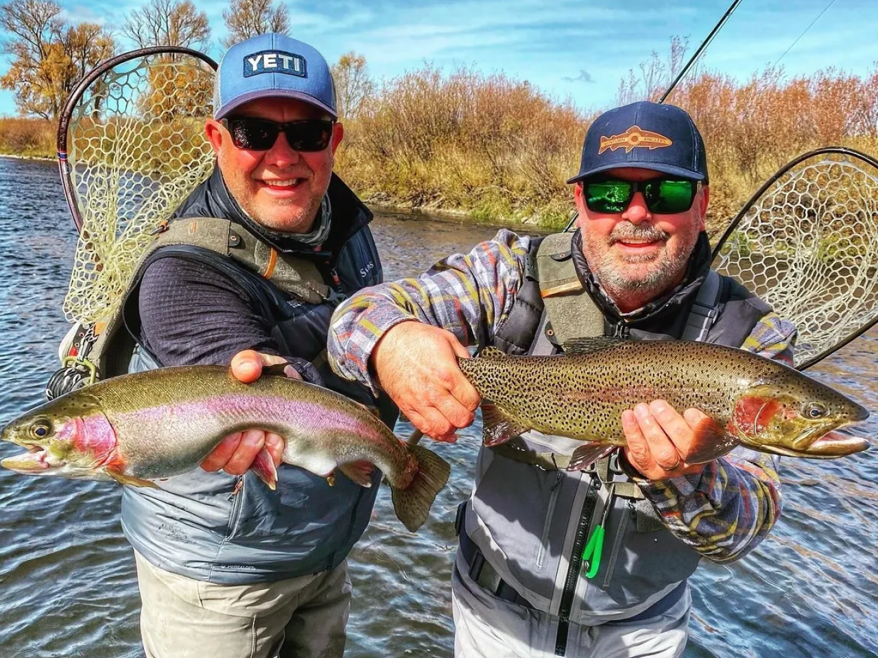 Two men holding up fish while standing on a river.