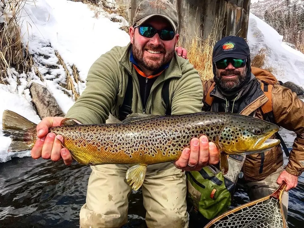 Two men holding a brown fish while standing on rocks.