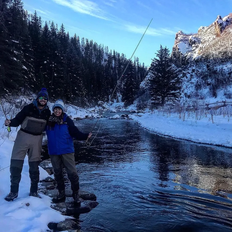 Two people standing on a river bank in the snow.